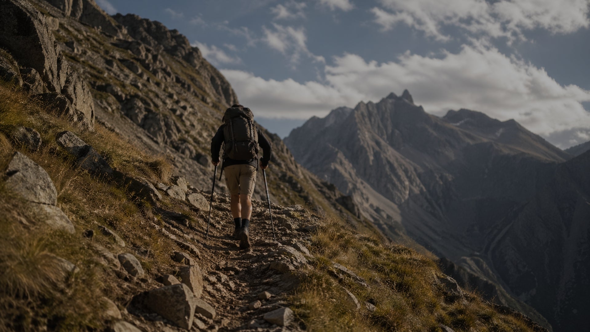 Hiker with a backpack and walking sticks ascending a mountain trail with dramatic mountain peaks in the background.